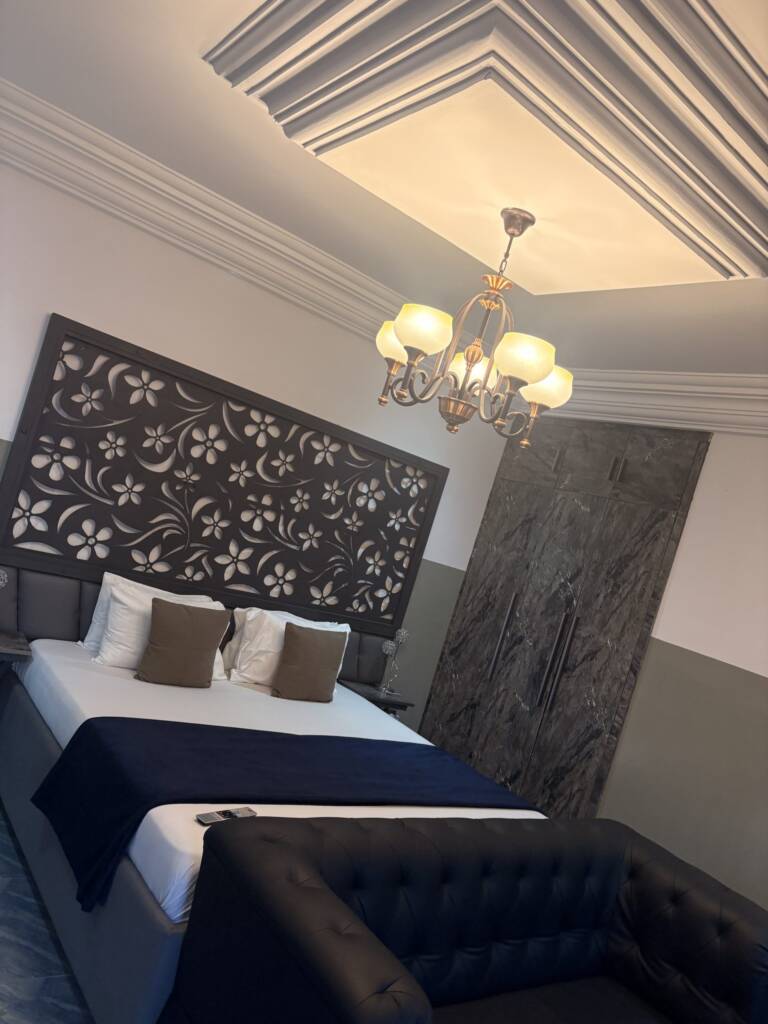 A guest room with navy bedding, a carved floral headboard panel and a chandelier